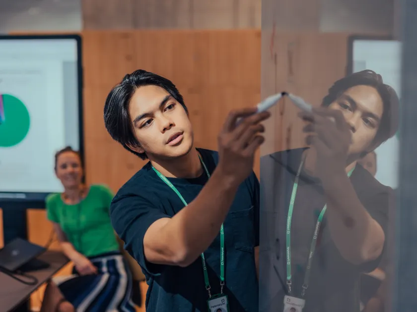 Patentstyrets ansatte samarbeider ved en whiteboard, der en person skriver med tusj mens en kollega følger med fra sidelinjen. I bakgrunnen vises en skjerm med et fargerikt sektordiagram.