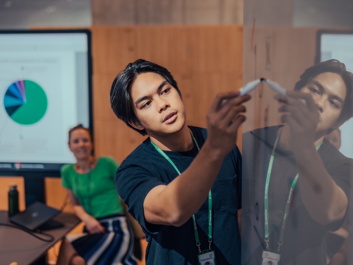 Patentstyrets ansatte samarbeider ved en whiteboard, der en person skriver med tusj mens en kollega følger med fra sidelinjen. I bakgrunnen vises en skjerm med et fargerikt sektordiagram.