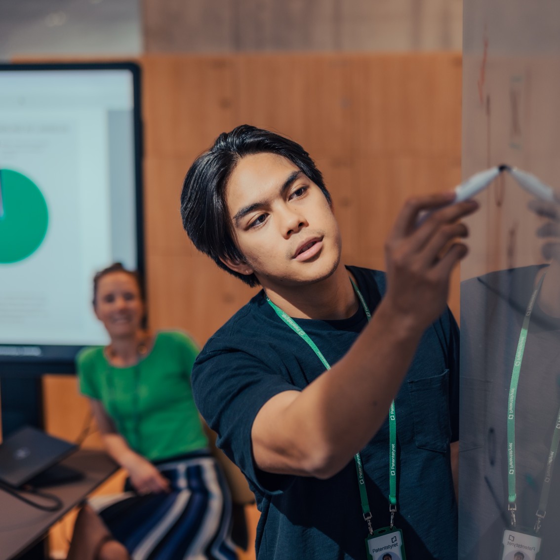 NIPO's employees collaborate at a whiteboard, where one person writes with a marker while a colleague watches from the sidelines. In the background, a screen with a colorful pie chart is displayed.