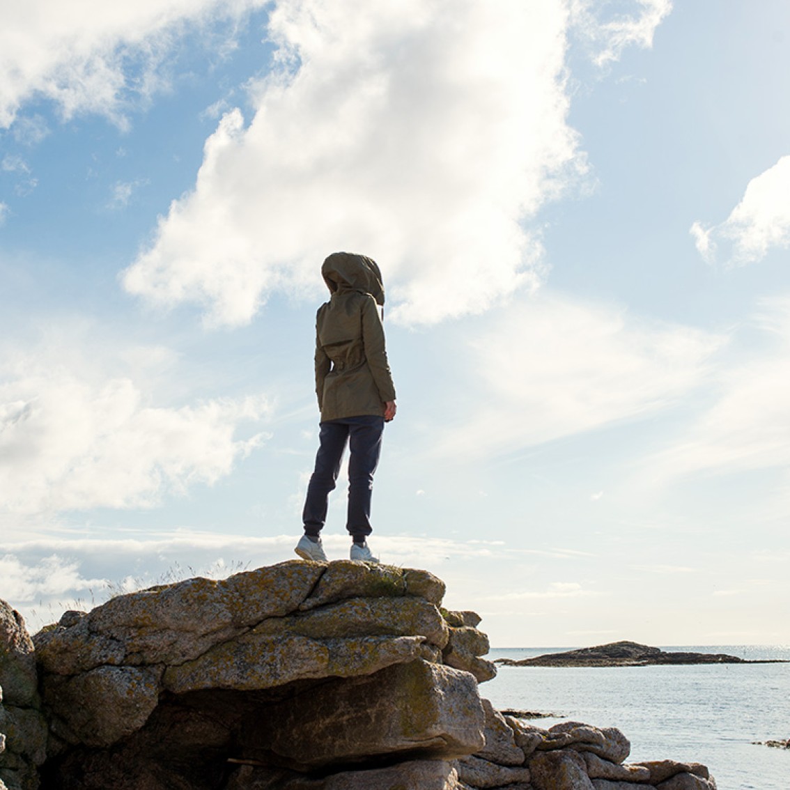 A person with a hood pulled over their head stands at the edge of a rocky shore, looking out over the sea.