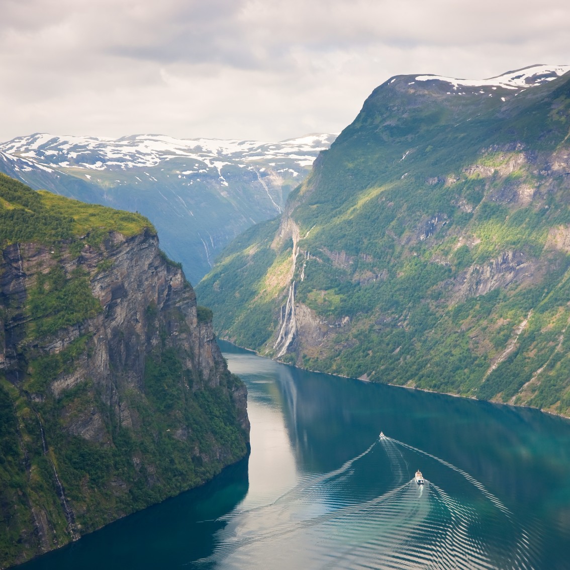 Landscape image of a Norwegian fjord surrounded by high, steep mountain slopes with green trees and snow-capped mountain peaks in the background. A boat makes wave patterns in the clear, blue water in the middle of the fjord. Cloudy sky gives soft light to the scenery.