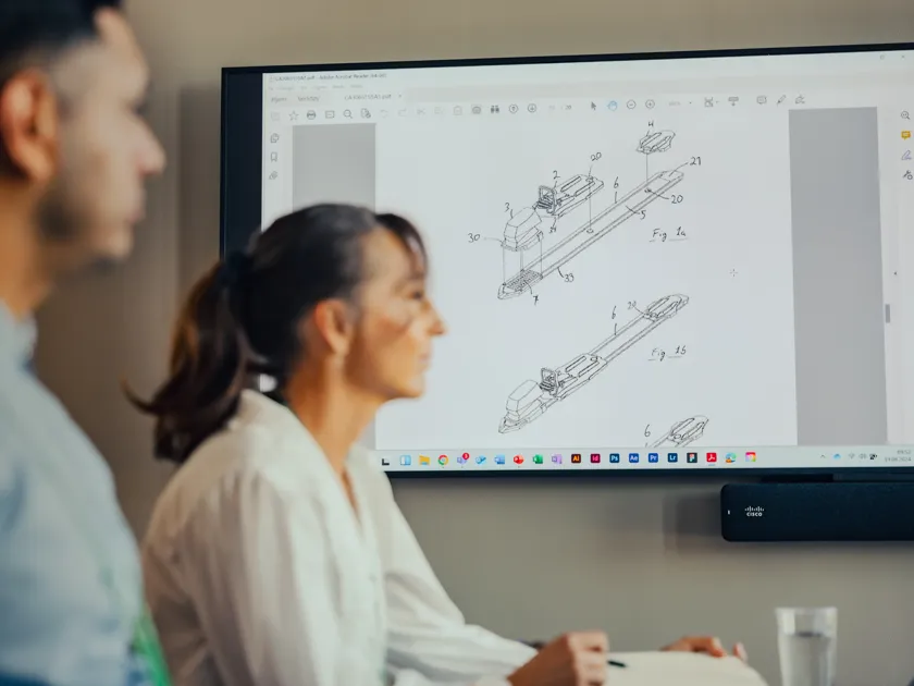 Two people in a meeting room with patent drawings showing on the big screen behind them