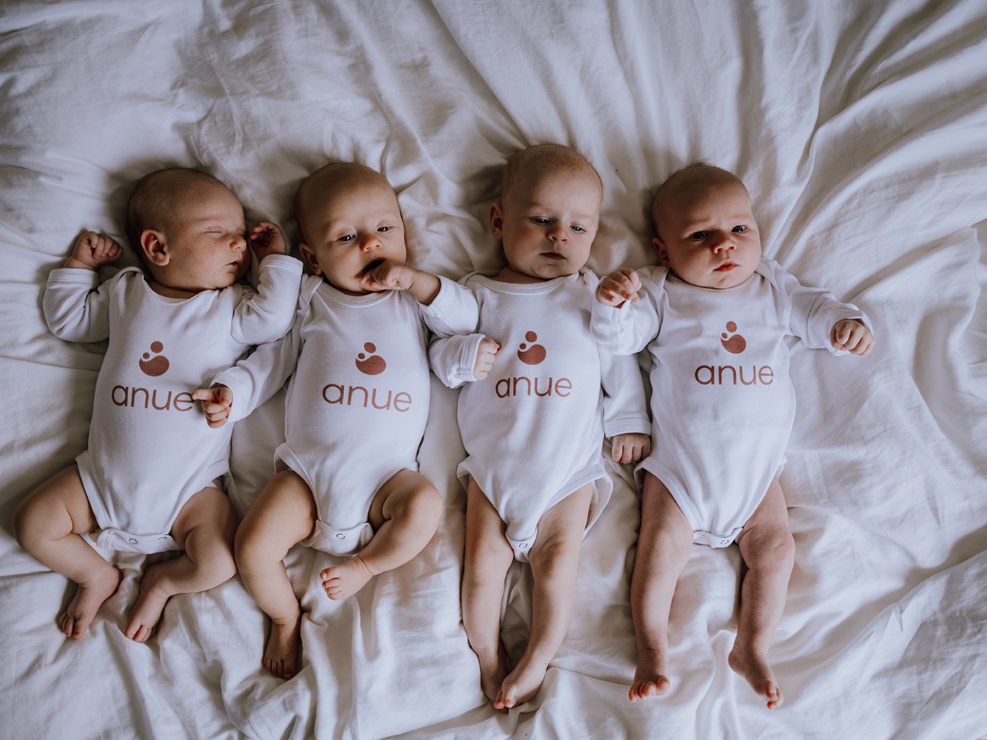 Fire babyer i en seng med bodyer merket med Anue