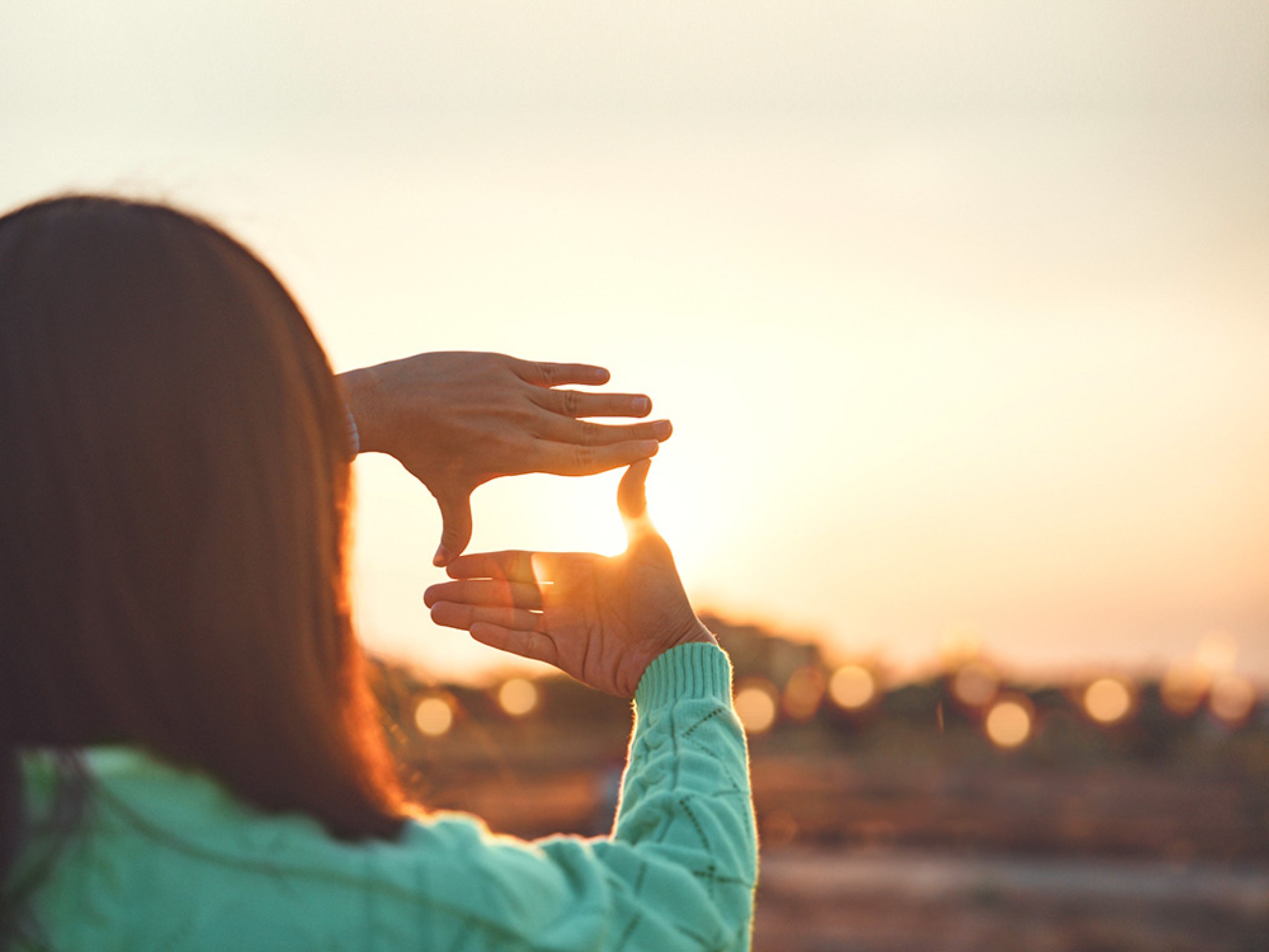 En kvinne lager en firkantet ramme med hendene rundt den sterke sola i et uskarpt landskap.